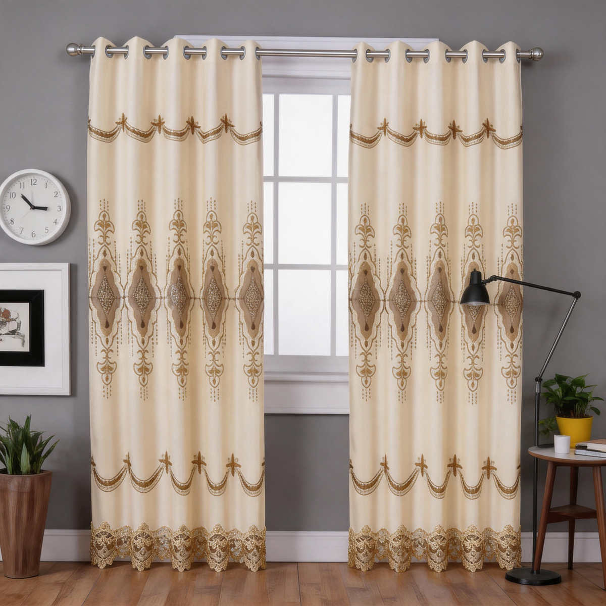 A pair of cream-colored grommet curtains with an ornate brown geometric and scroll pattern, hanging on a silver rod in a styled room setting with a window, wall clock, lamp, and side table.