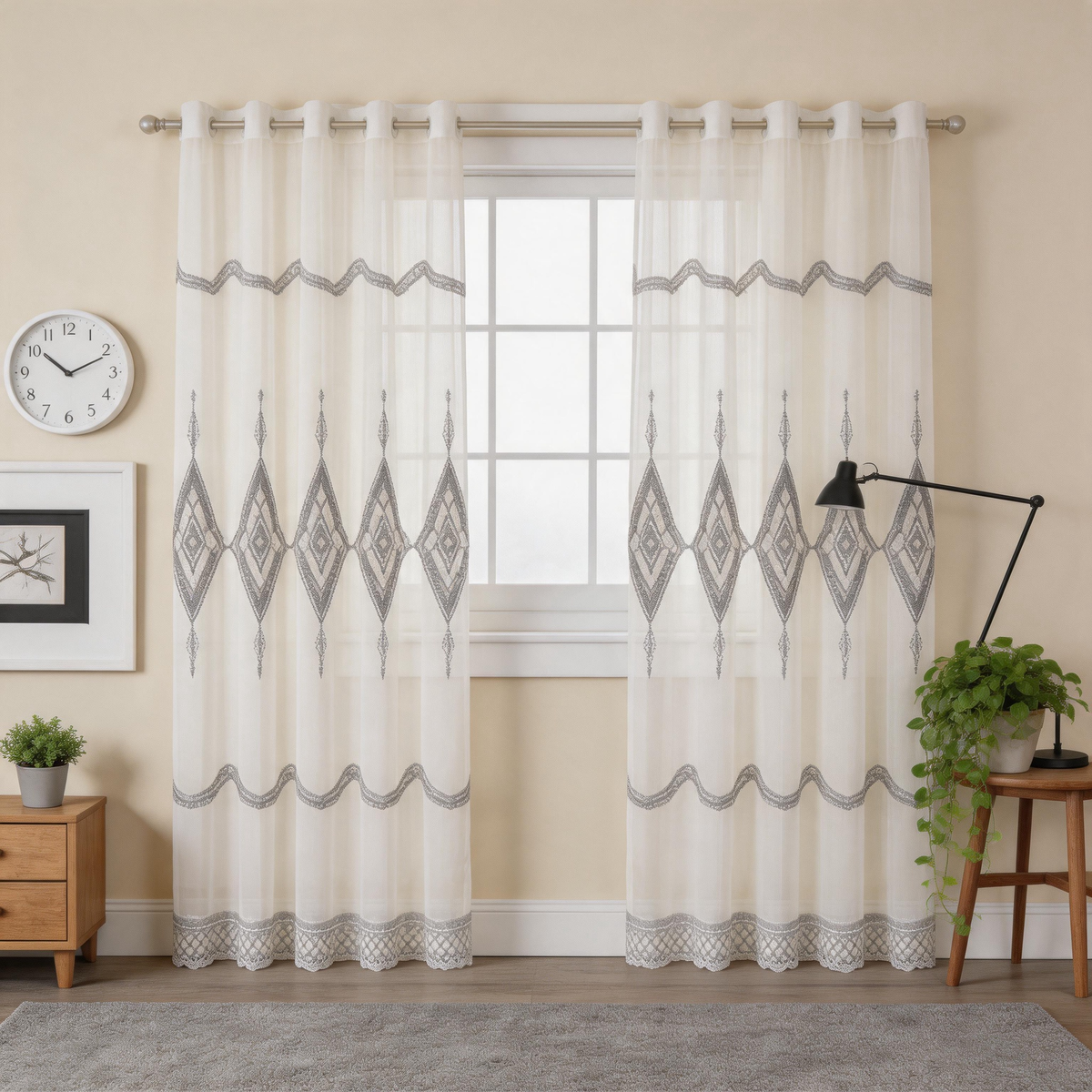 A pair of sheer off-white curtain panels with gray geometric diamond and wavy pattern details, hanging on a white rod in a styled room setting with a window, side table, lamp, and plants.